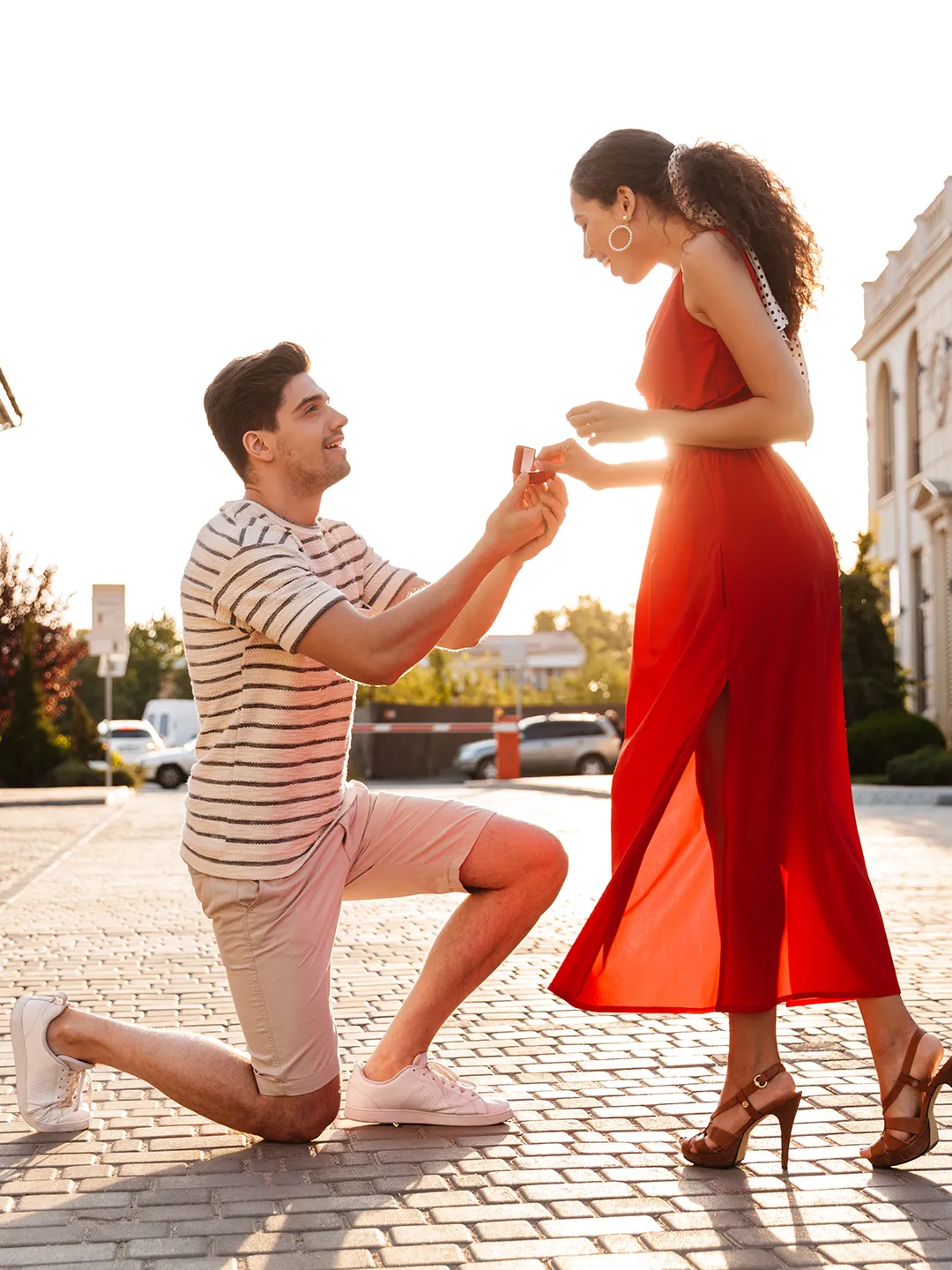 A man in a striped shirt kneels on one knee while presenting an engagement ring to a woman wearing a red dress in an outdoor urban plaza during golden hour.