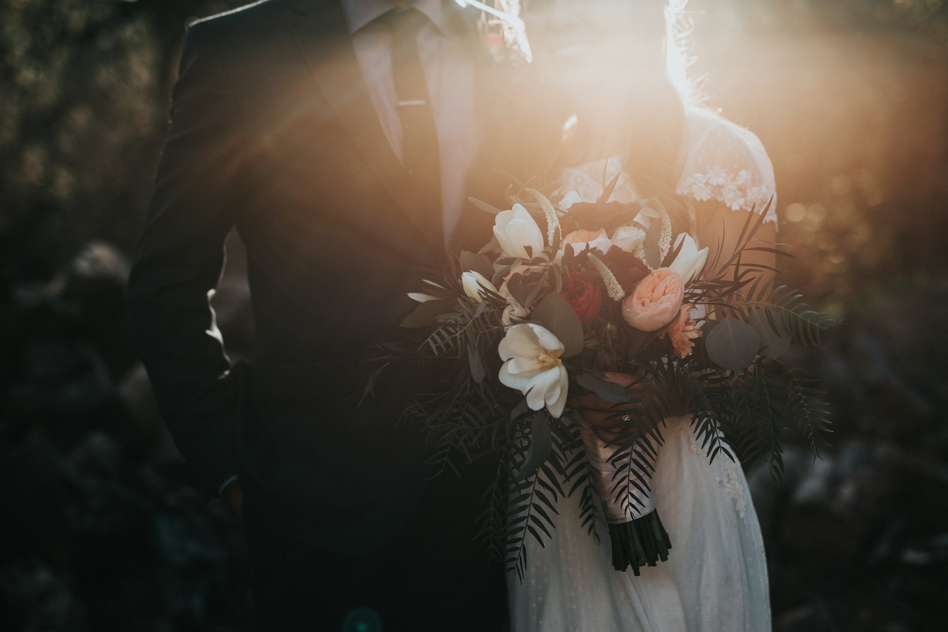 A bride in a white lace dress holds a romantic wedding bouquet of white flowers, peach roses, and green ferns, backlit by golden sunlight during an outdoor ceremony.